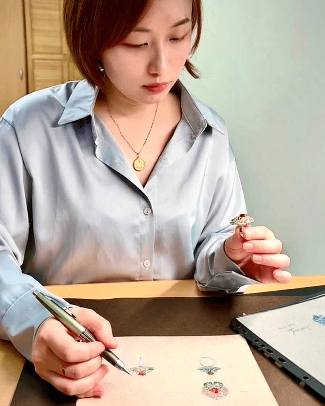 Tru-Mi 設計師 Jessica 正在繪製客製珠寶設計草圖，手中拿著彩寶戒指作品。